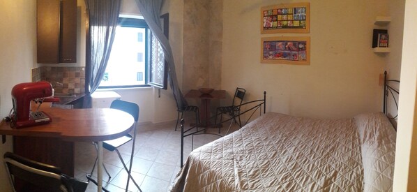 A small, compact room with a bed, a dining table with chairs, a kitchenette, and a window with curtains.