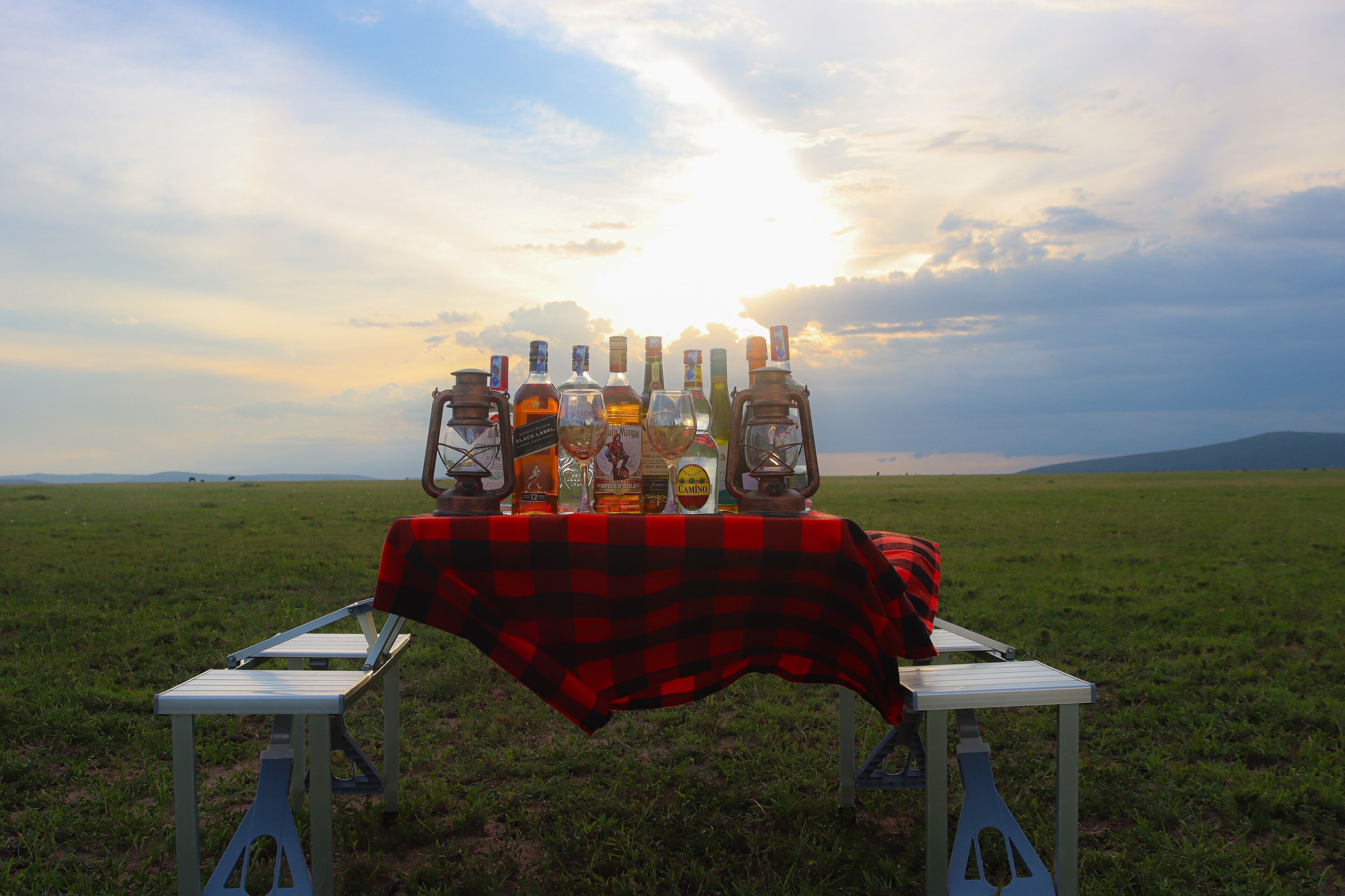 A picnic setup with a table covered by a red and black checkered tablecloth, surrounded by bottles and lanterns, set against a sunset over a grassy field.