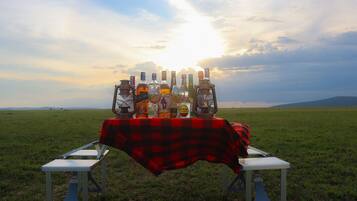 A picnic setup with a table covered by a red and black checkered tablecloth, surrounded by bottles and lanterns, set against a sunset over a grassy field.
