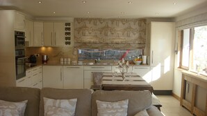A modern kitchen with white cabinets, a dining area with a table set for six, and a living room with a beige sofa and remote controls on a coffee table.