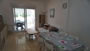 Un coin repas avec une table recouverte d’une nappe, des chaises blanches et un banc. Une porte-fenêtre coulissante donne sur un espace extérieur aménagé avec des chaises et une table. On peut voir une horloge et un tableau accrochés au mur.
