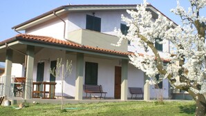A two-story house with a covered porch, two benches, and a flowering tree. - Bright country villa, with veranda, pool, garden, near Florence (Buggiano)