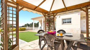 Eine überdachte Terrasse mit Tisch und Stühlen, ein Holz-Pergola und Blick auf ein Haus und einen Rasen.