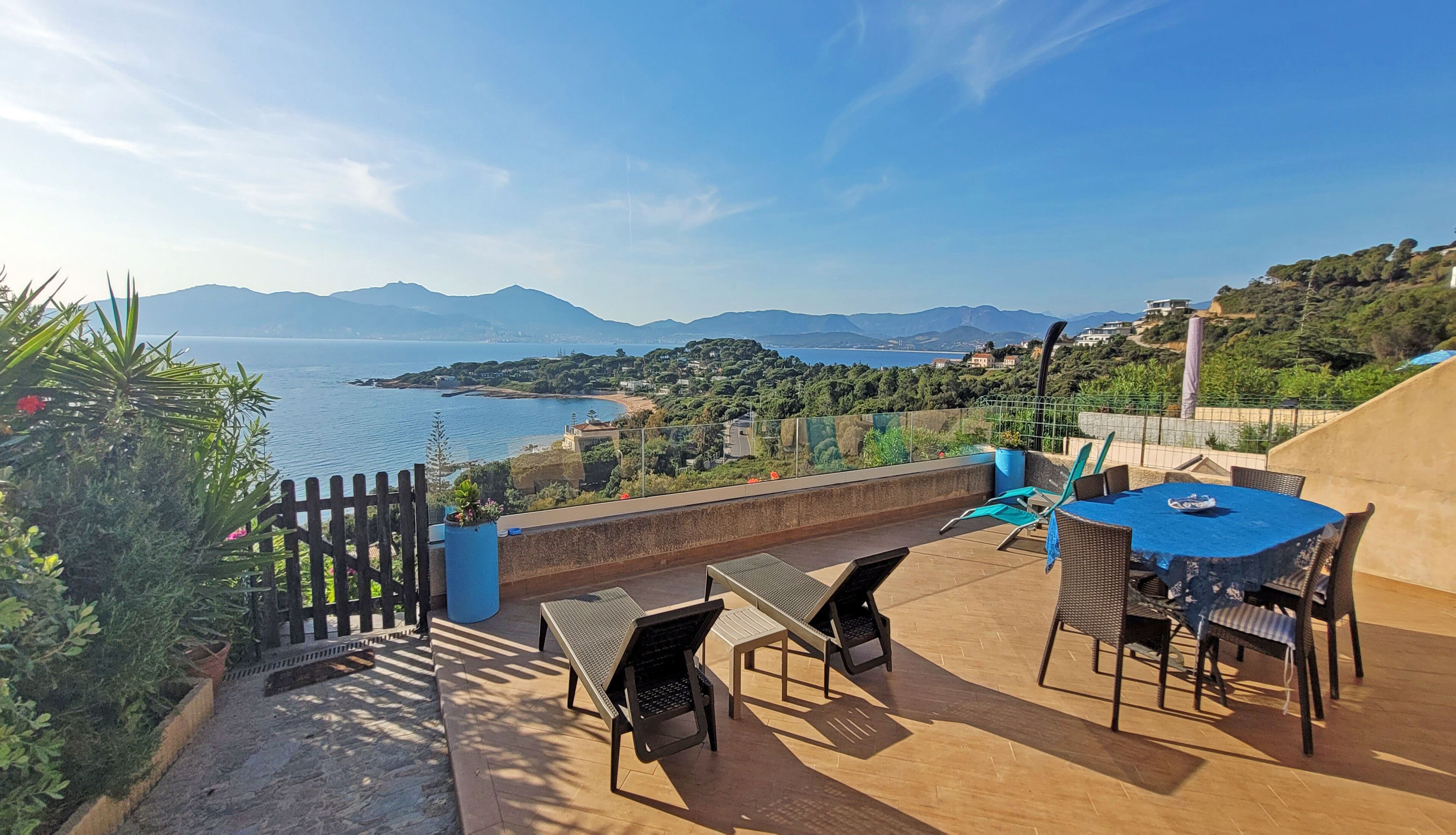 An outdoor terrace with a table and chairs, overlooking a coastal landscape with mountains in the distance.