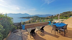Una terraza al aire libre con una mesa y sillas, con vista a un paisaje costero con montañas a lo lejos.