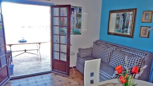 Una sala de estar con un sofá a cuadros, una mesa de comedor con mantel floral y una puerta que da a un área exterior con mesa y sillas.