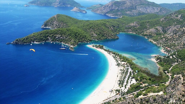 Une vue aérienne d’une zone côtière avec une plage, des eaux d’un bleu limpide et des collines verdoyantes.