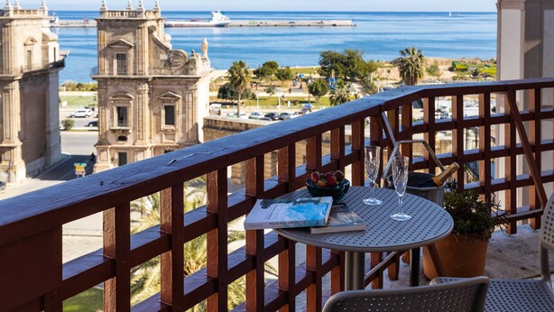 Uma varanda com vista para o oceano e um edifício histórico. Há uma mesa com um livro, uma tigela de frutas e uma garrafa de champanhe.