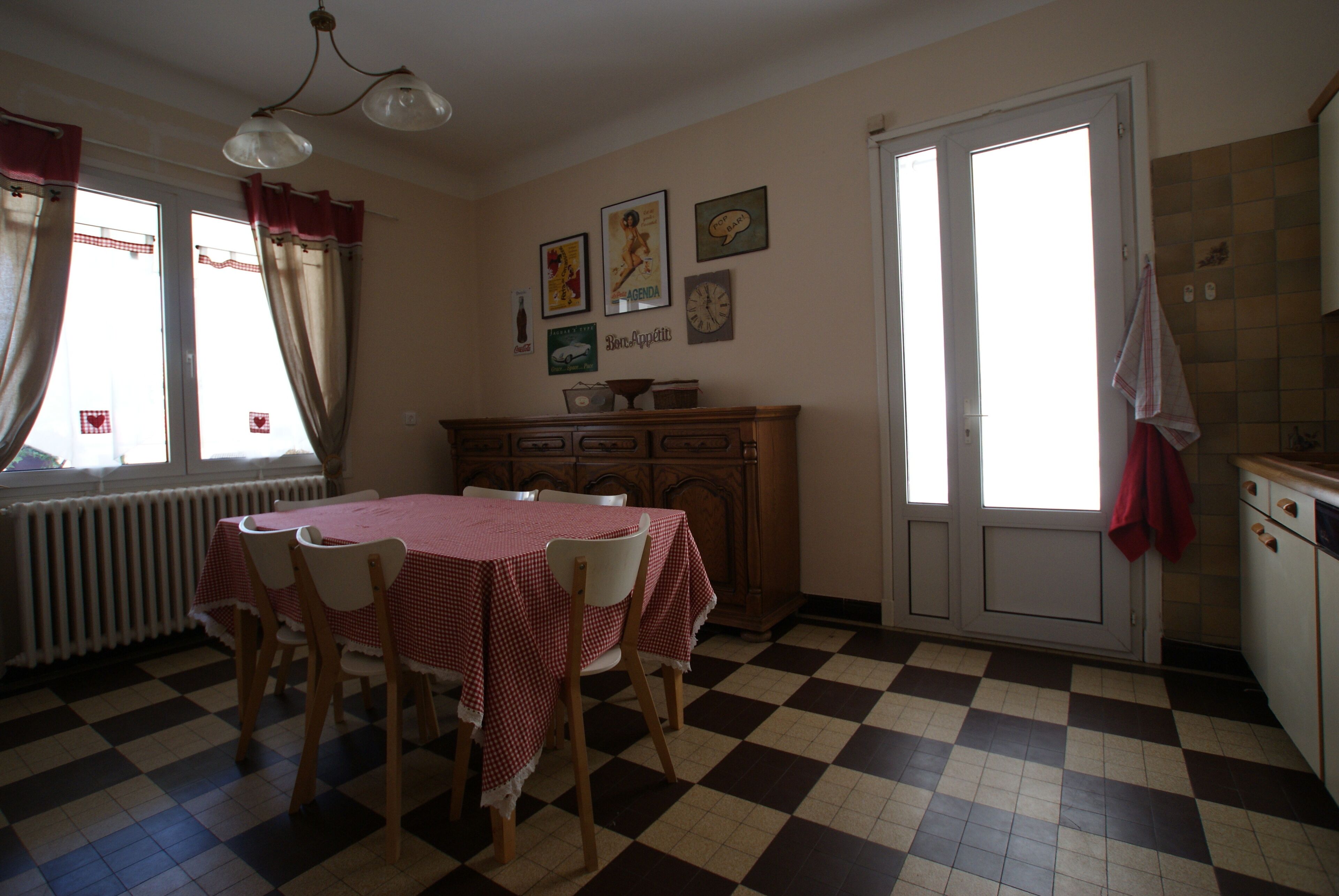 Ein Essbereich mit kariertem Boden, einem Tisch mit rotem Tischläufer und Stühlen. An der Wand hängen Bilder und in der Nähe des Fensters steht eine Heizung.