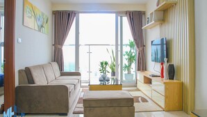 Ein Wohnzimmer mit einer Couch, einem Hocker und einem Fernseher auf einem Holzboden. Ein Glastisch und ein Blumentopf sind ebenfalls vorhanden.