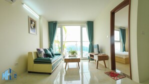 Ein Wohnzimmer mit Sofa, Couchtisch, Schreibtisch mit Computer und einem Bett. Ein Fenster mit Vorhängen, eine Pflanze und Blick auf Gebäude sind ebenfalls zu sehen.