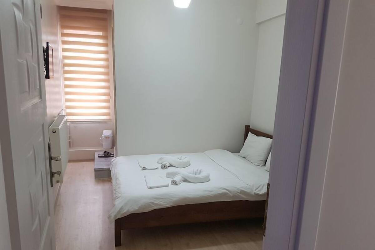 Ein Schlafzimmer mit einem hölzernen Bett, weißen Bettwäsche, einem Nachttisch mit Lampe und einem Fenster mit Jalousien.