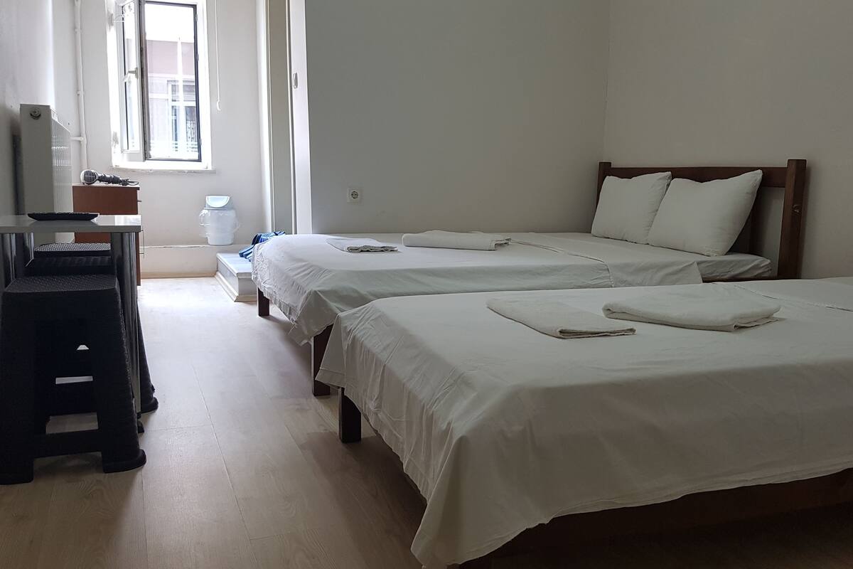Ein Hotelzimmer mit zwei Einzelbetten, einem kleinen Schreibtisch mit Stuhl, einem Heizkörper und einem Fenster mit Vorhängen.