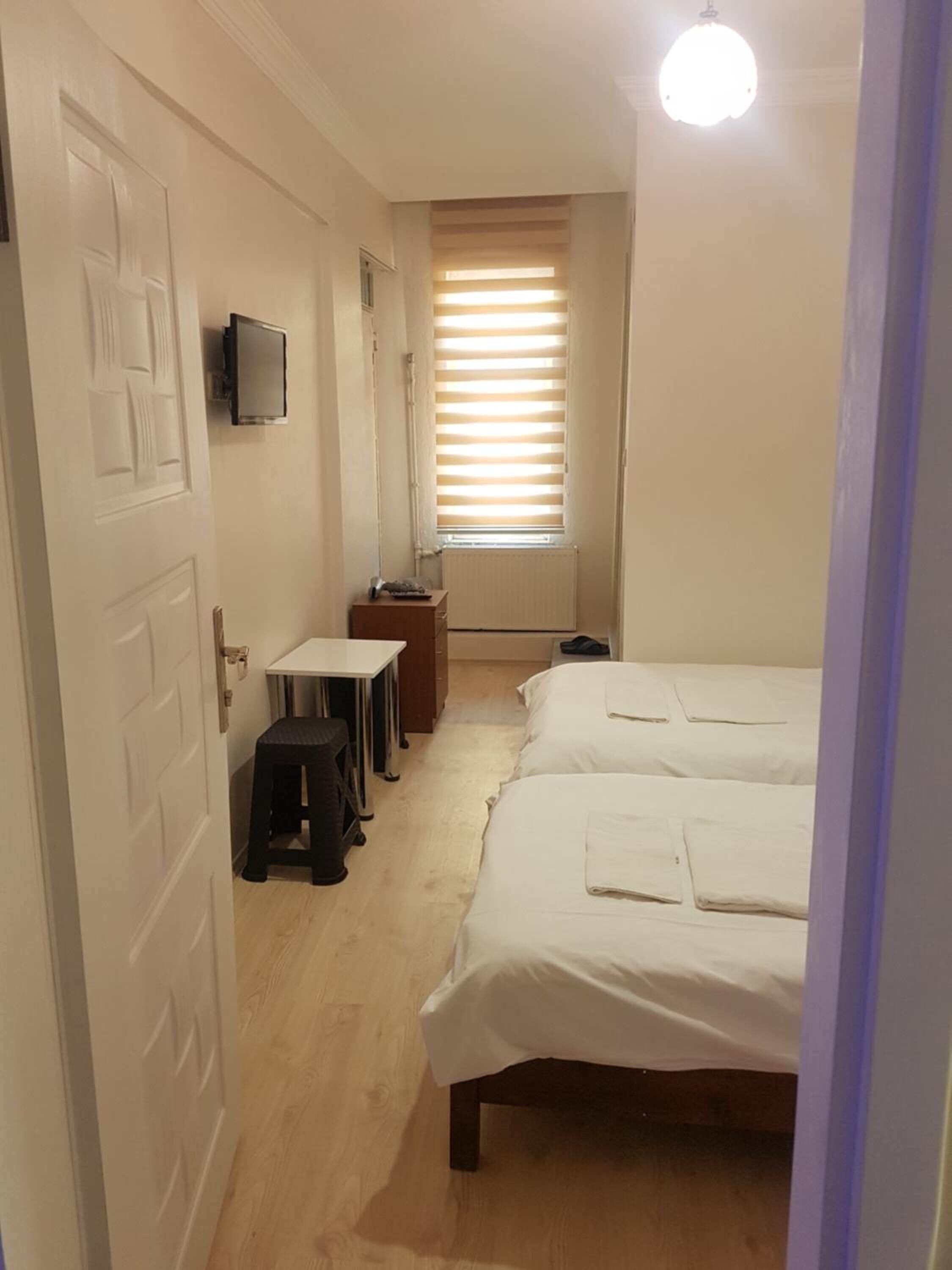 Ein schmales Hotelzimmer mit zwei Betten, einem kleinen Tisch, einem Hocker, einem an der Wand montierten Fernseher und einem Fenster mit Jalousien.