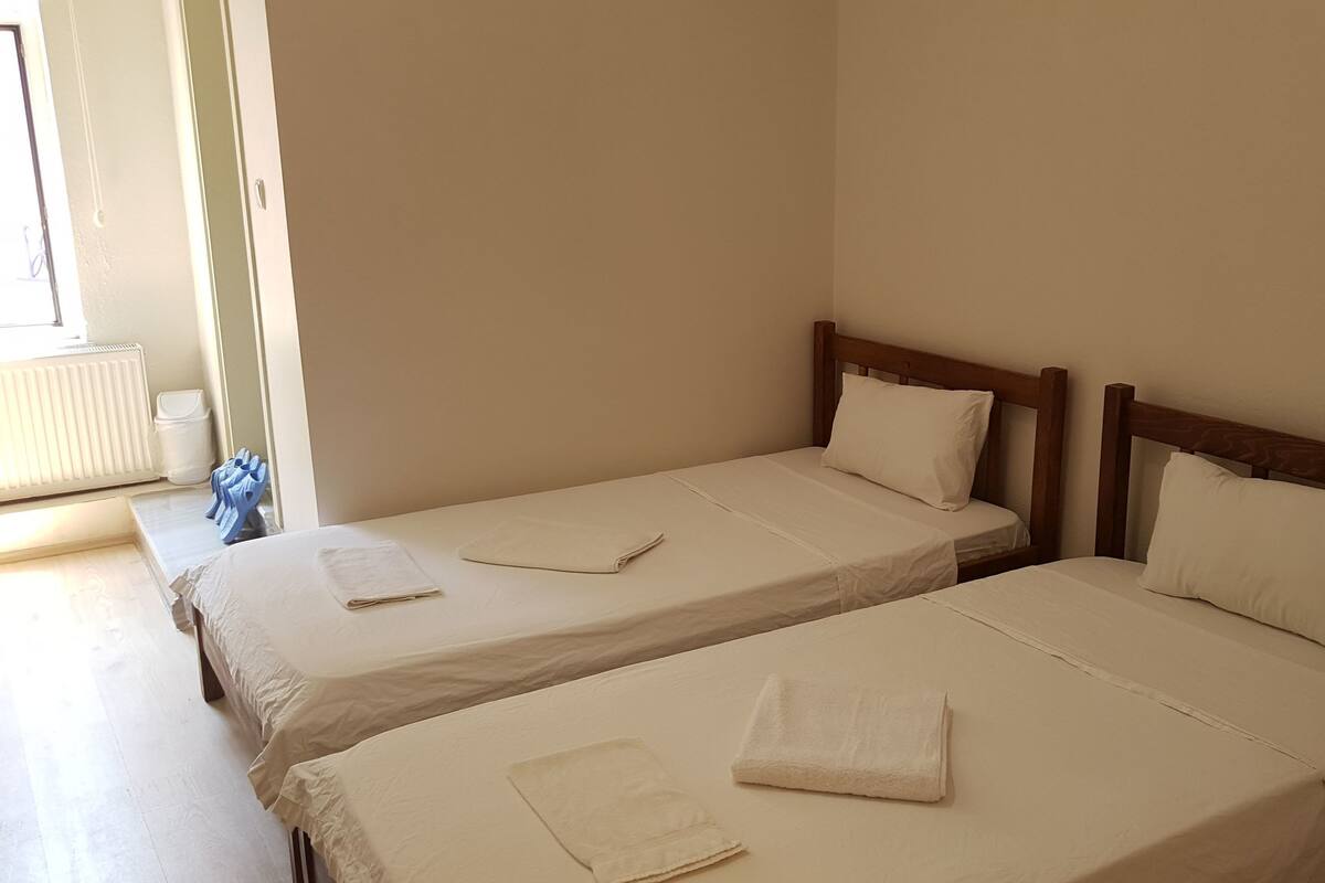 Ein Zimmer mit zwei Einzelbetten, jeweils mit einem hölzernen Kopfteil und weißer Bettwäsche. Eine Heizung und ein Mülleimer sind in der Ecke sichtbar.