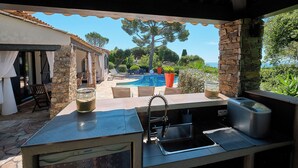 An outdoor kitchen area with a stone wall, a pool, and a view of the sea. - 400m from the sea, Villa with splendid sea view, heated infinity pool (Fréjus)