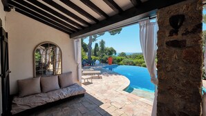 A room with a stone wall, a window with a view of a pool and greenery, and a cushioned bench. - 400m from the sea, Villa with splendid sea view, heated infinity pool (Fréjus)