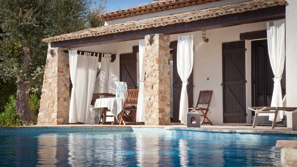 Un espace aménagé au bord de la piscine, avec un mur en pierre, deux chaises en bois, une table et un coin salon couvert par des rideaux.