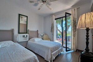 Une chambre d’hôtel avec deux lits, un ventilateur de plafond, un miroir, une lampe et une porte-fenêtre coulissante donnant sur un espace extérieur.