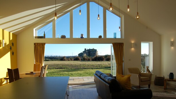 A spacious living area with a large window overlooking a grassy field and a distant building. There is a dining table and a sofa with cushions.