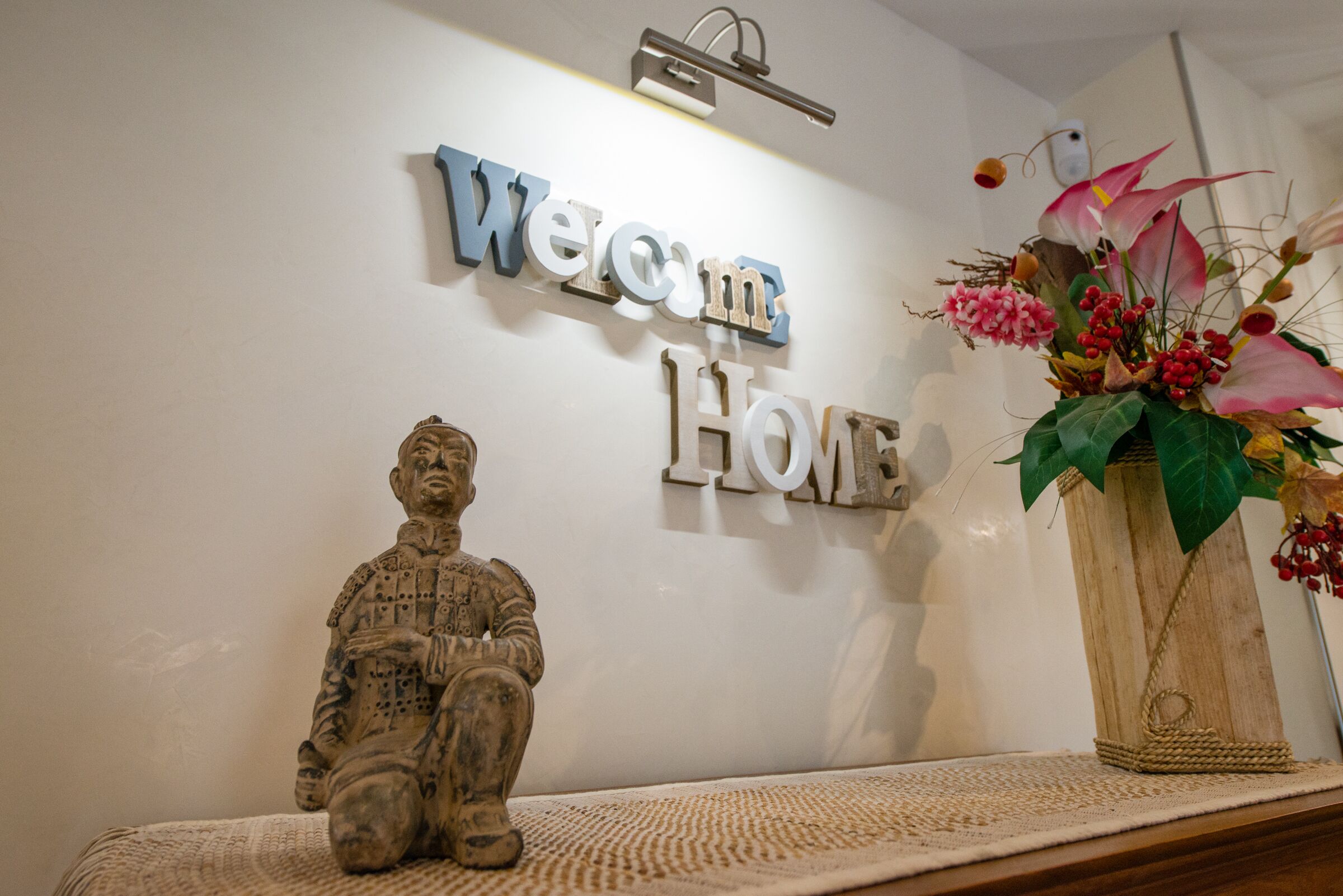 Ein Willkommensschild mit der Aufschrift 'Welcome Home' in einem Zimmer mit einer dekorativen Statue und einem Blumenstrauß.