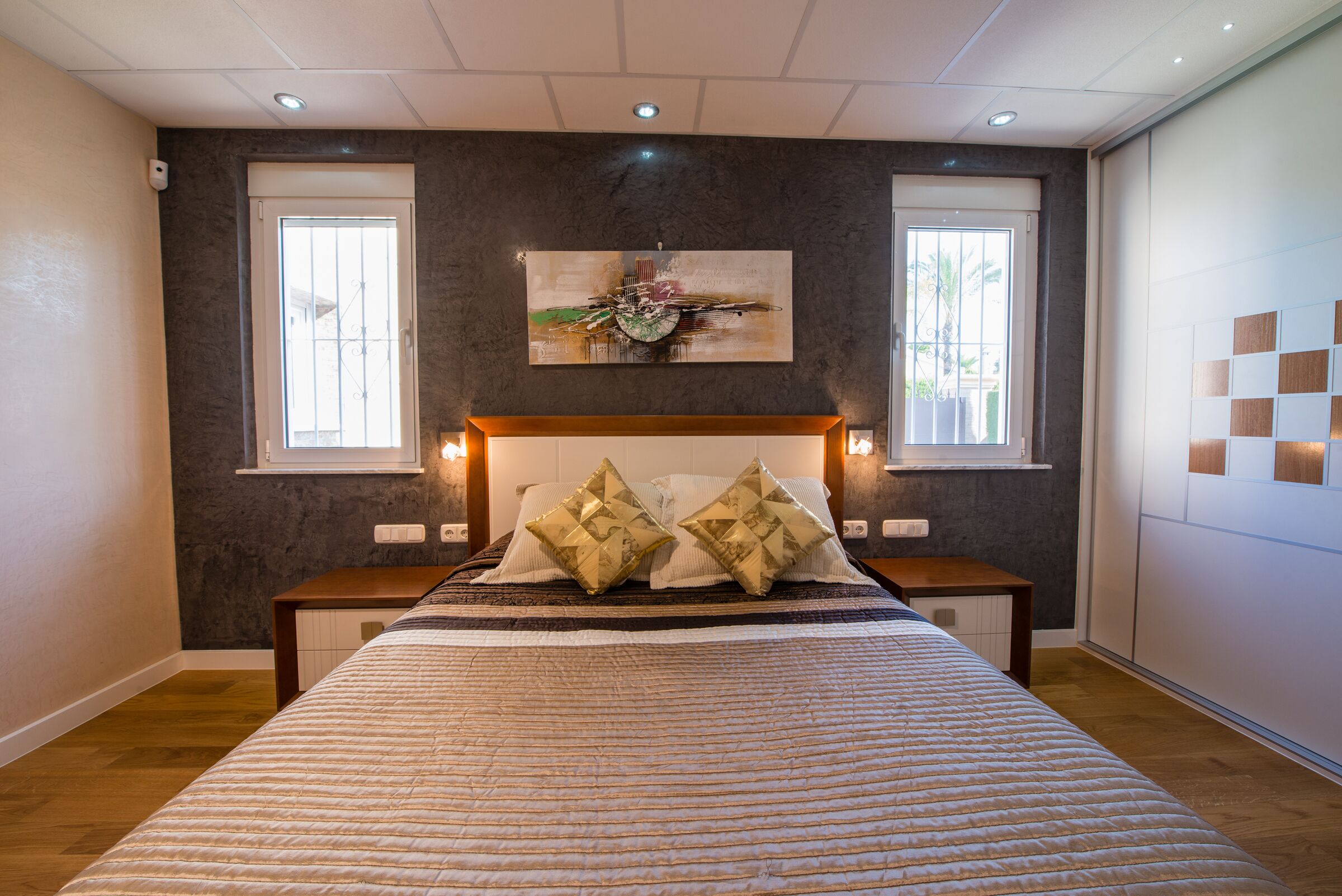 Quarto com uma cama grande, duas mesas de cabeceira, uma cabeceira, um quadro pendurado na parede e duas janelas com cortinas.