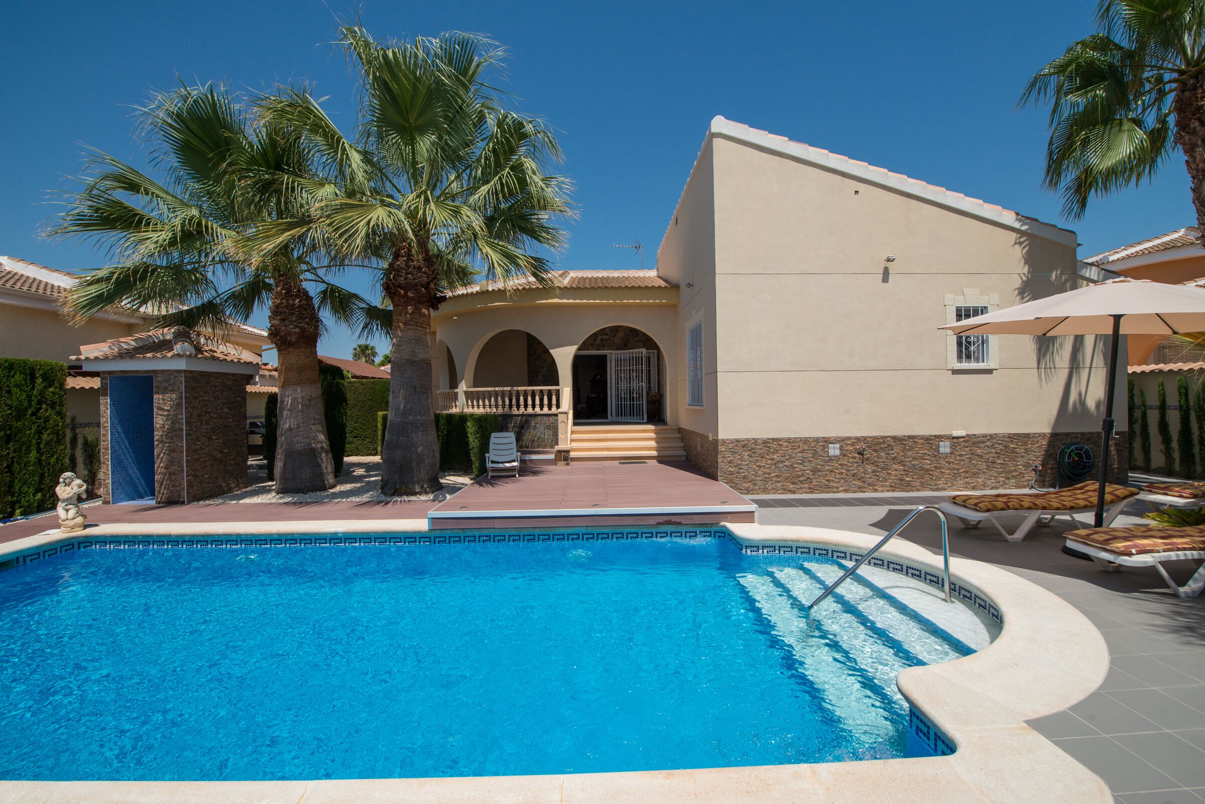 A swimming pool with palm trees, a covered patio, and a building with a tiled roof.