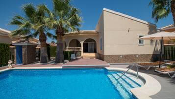 A swimming pool with palm trees, a covered patio, and a building with a tiled roof.