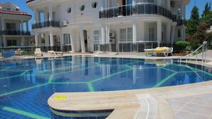 Una piscina con solárium empedrado, sillones de descanso y un edificio con balcones al fondo.
