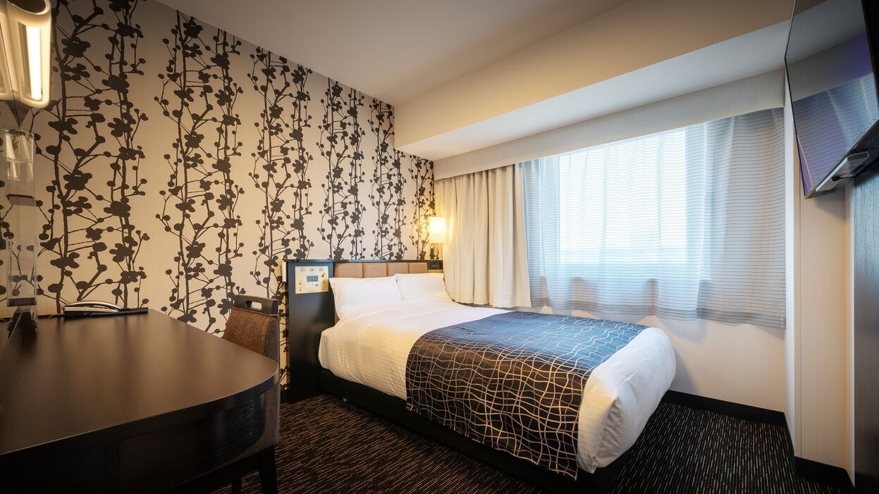 A hotel room with a bed, a desk, a chair, a TV, and a window with curtains.