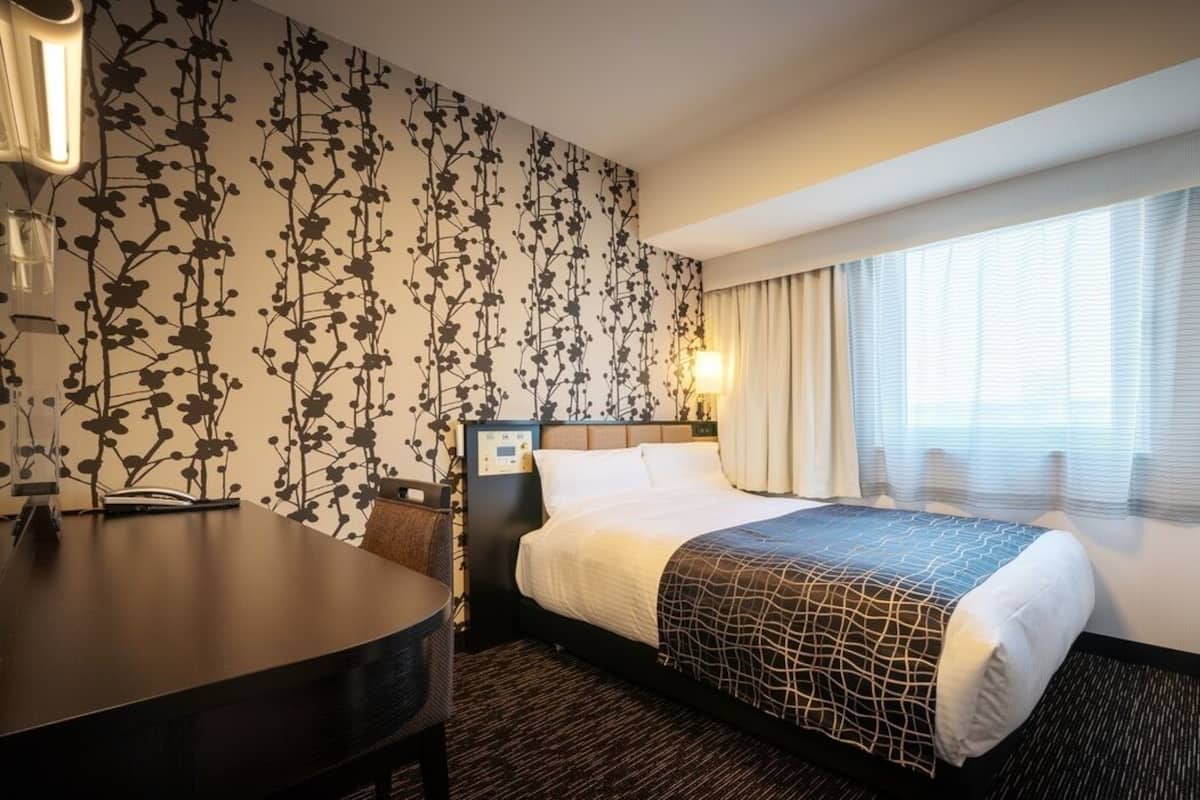Habitación de hotel con cama, escritorio, silla, televisor y ventana con cortinas.