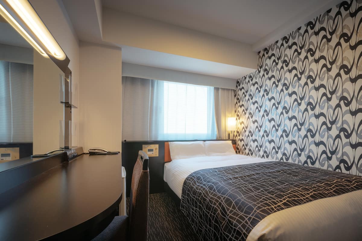 Habitación de hotel con cama, escritorio, silla, espejo, ventana con cortinas y pared con diseño.