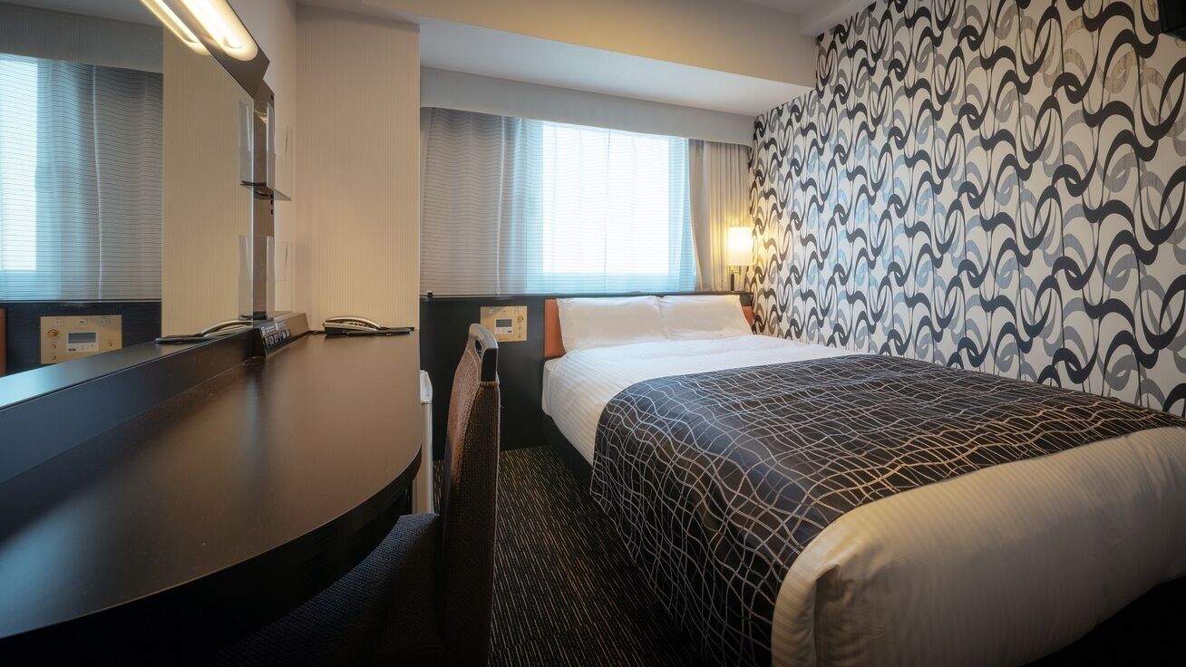 A hotel room with a bed, a desk with a telephone, a chair, a window with curtains, and a patterned wall.