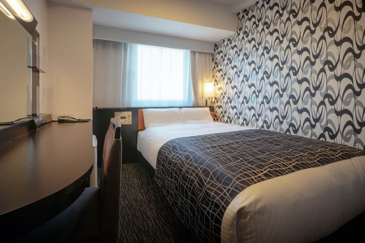 Una habitación de hotel con cama, escritorio con teléfono, silla, ventana con cortinas y pared con diseño.