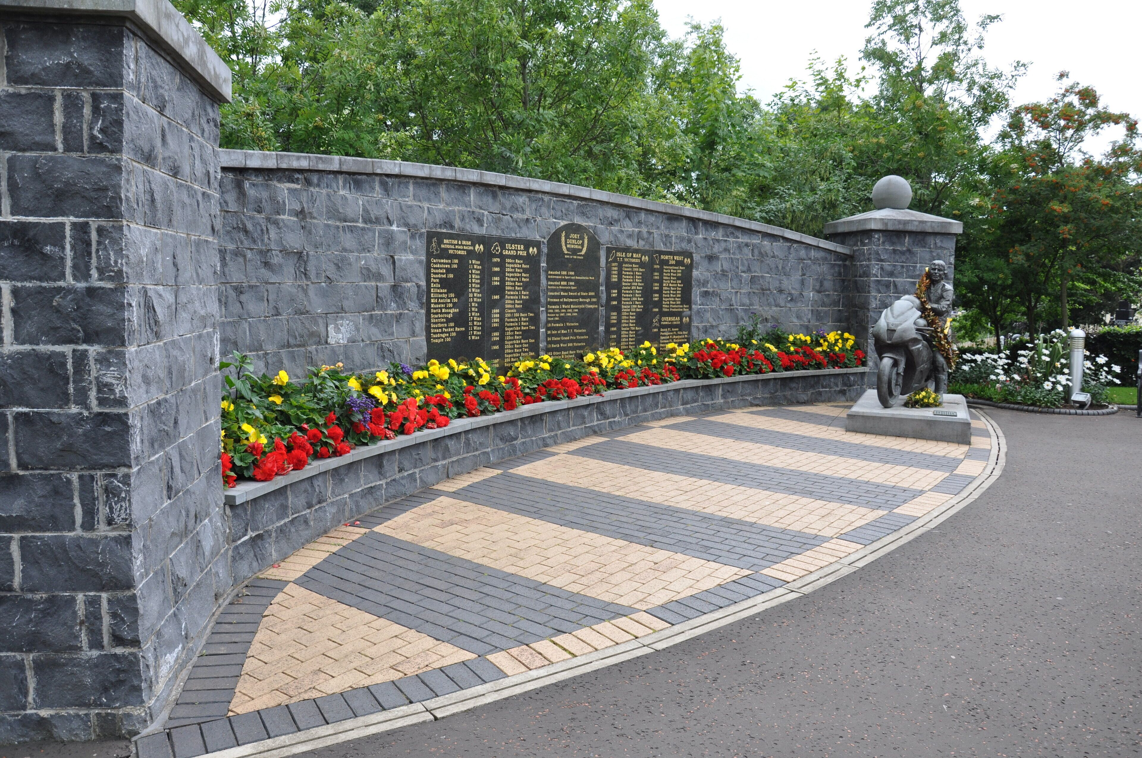 A stone wall with engraved plaques, a statue, and a flower bed.