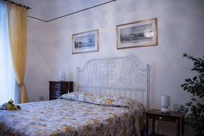 A bedroom with a bed, two framed pictures on the wall, a bedside table with a lamp, and a plant.
