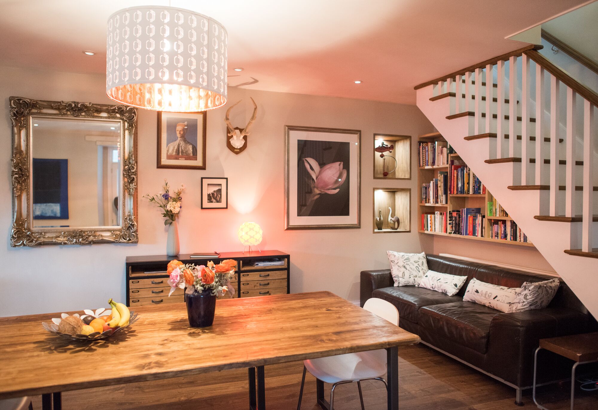 Ein Wohnzimmer mit einem Holzesstisch, einem braunen Ledersofa, einer Treppe und einem Bücherregal.