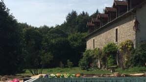 A swimming pool with clear water, surrounded by greenery and a stone building with a tiled roof.