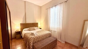Un dormitorio con cama, lámparas de noche, un banco de mimbre y una ventana con cortinas.