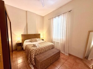 A bedroom with a bed, bedside lamps, a wicker bench, and a window with curtains.