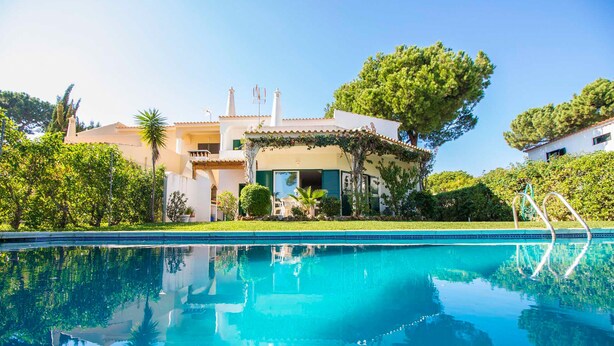 Una piscina con un reflejo claro, una casa con techo de tejas y un árbol grande al fondo.