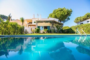 A pool with a clear reflection, a house with a tiled roof, and a large tree in the background.