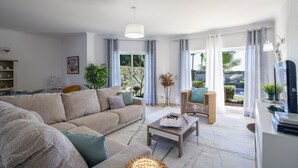 Una sala moderna con un sofá beige, una mesa de centro y una silla de mimbre. Hay un ventanal con cortinas, una planta y vistas al exterior.