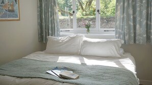 Una cama bien tendida con sábanas blancas, un libro sobre la cama y una ventana con cortinas.