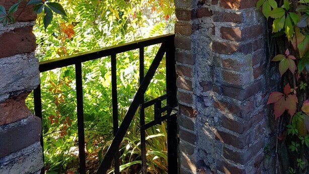 Una entrada de ladrillo en arco con una reja de metal, rodeada de follaje y árboles.