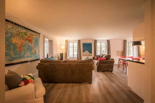 A spacious living room with a large world map, two sofas, a dining area with red chairs, and a view of a mountain through a window.