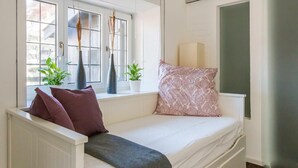 Um quarto pequeno e acolhedor com uma cama branca, uma janela com vista e uma mesa de cabeceira com abajur.