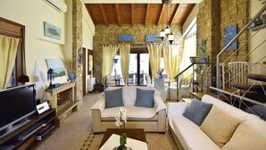 A spacious living area with a stone wall, wooden beams, a sofa set, a TV, and a staircase. - BEAUTIFUL SANI VILLA (Chalkidiki)