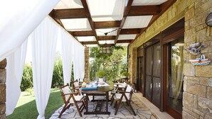 An outdoor patio with a stone floor, wooden table, and chairs, surrounded by a brick wall and a white canopy. - BEAUTIFUL SANI VILLA (Chalkidiki)