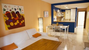 Una sala de estar moderna con un sofá blanco, una mesa de centro de madera y cojines naranjas. En el fondo, se ve un comedor con una mesa y sillas blancas.